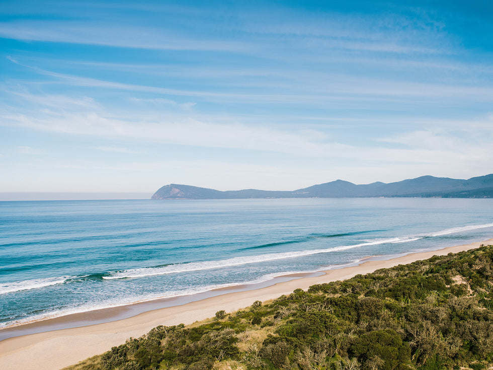 Australian sandy beach with ocean waves, lush green vegetation, and a mountain range in the background under a clear blue sky.